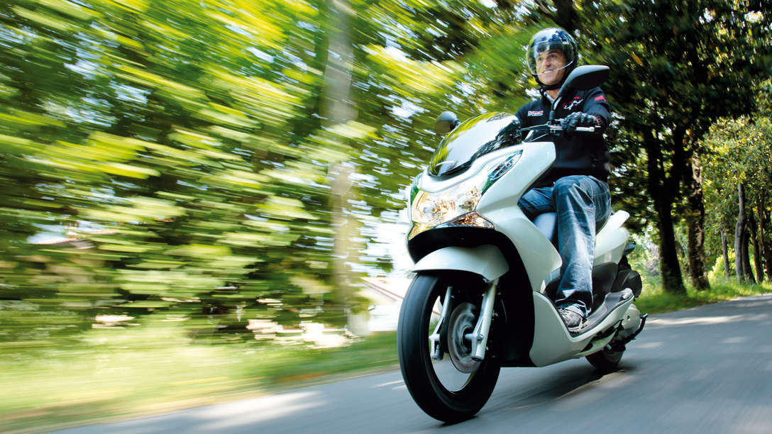 Egy Honda PCX elölről, motorossal, háromnegyedes nézetben. Bal oldali profil (közúton).