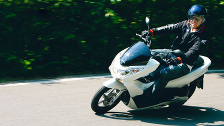Egy kanyarodó Honda PCX eleje, motorossal, háromnegyedes nézetben. Bal oldali profil (közúton).
