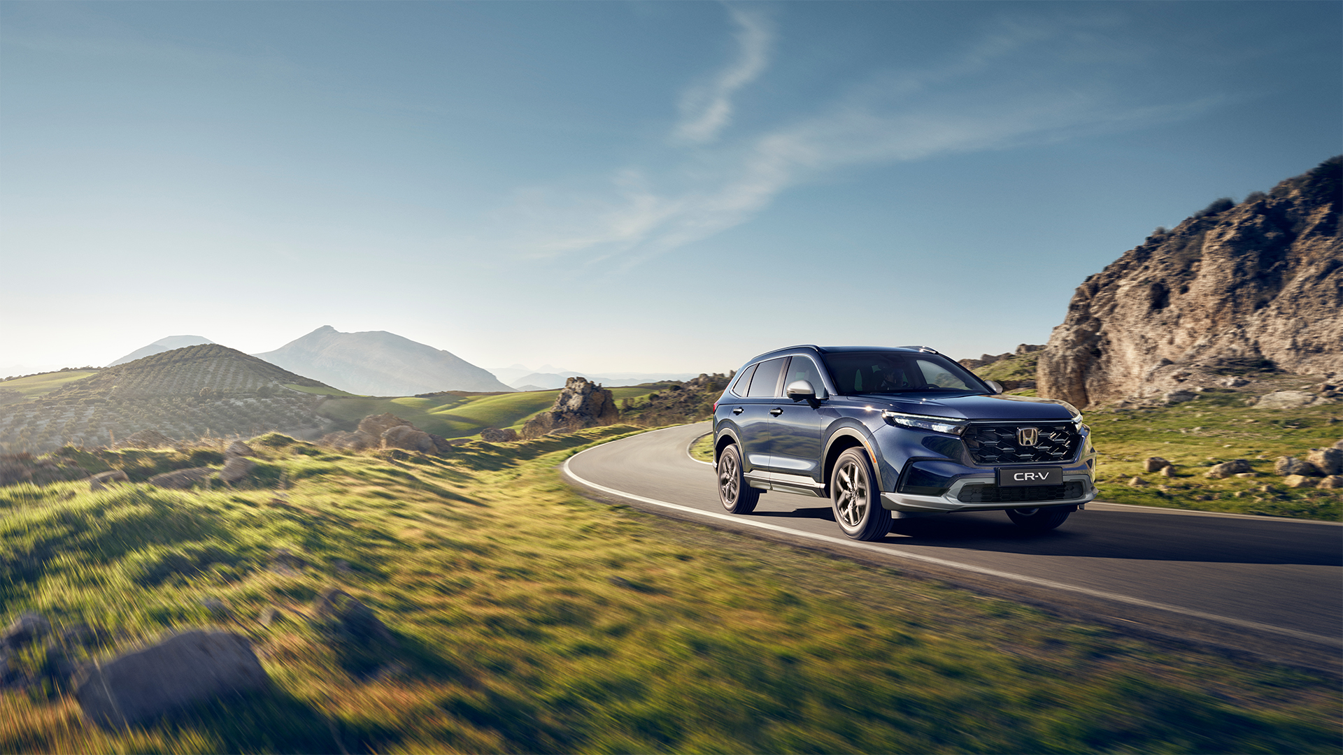 A dark blue Honda CR-V drives along a winding mountain road surrounded by lush green grass and rocky terrain. The landscape features rolling hills and distant mountains under a clear blue sky with wispy clouds.
