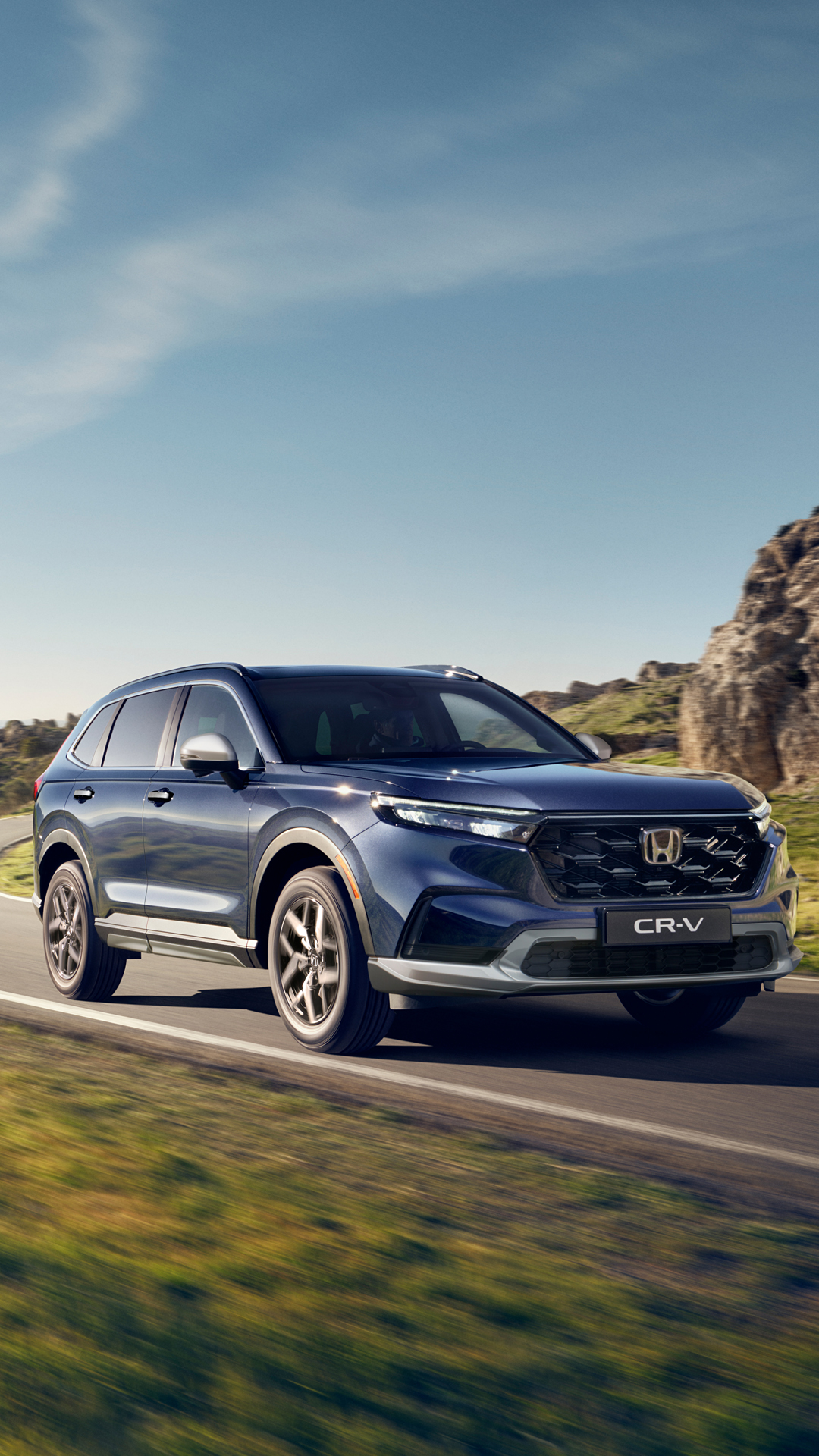 A blue Honda CR-V SUV driving on a road with rocky terrain and blue sky in the background. The vehicle is shown in motion, highlighting its sleek design and rugged capabilities.