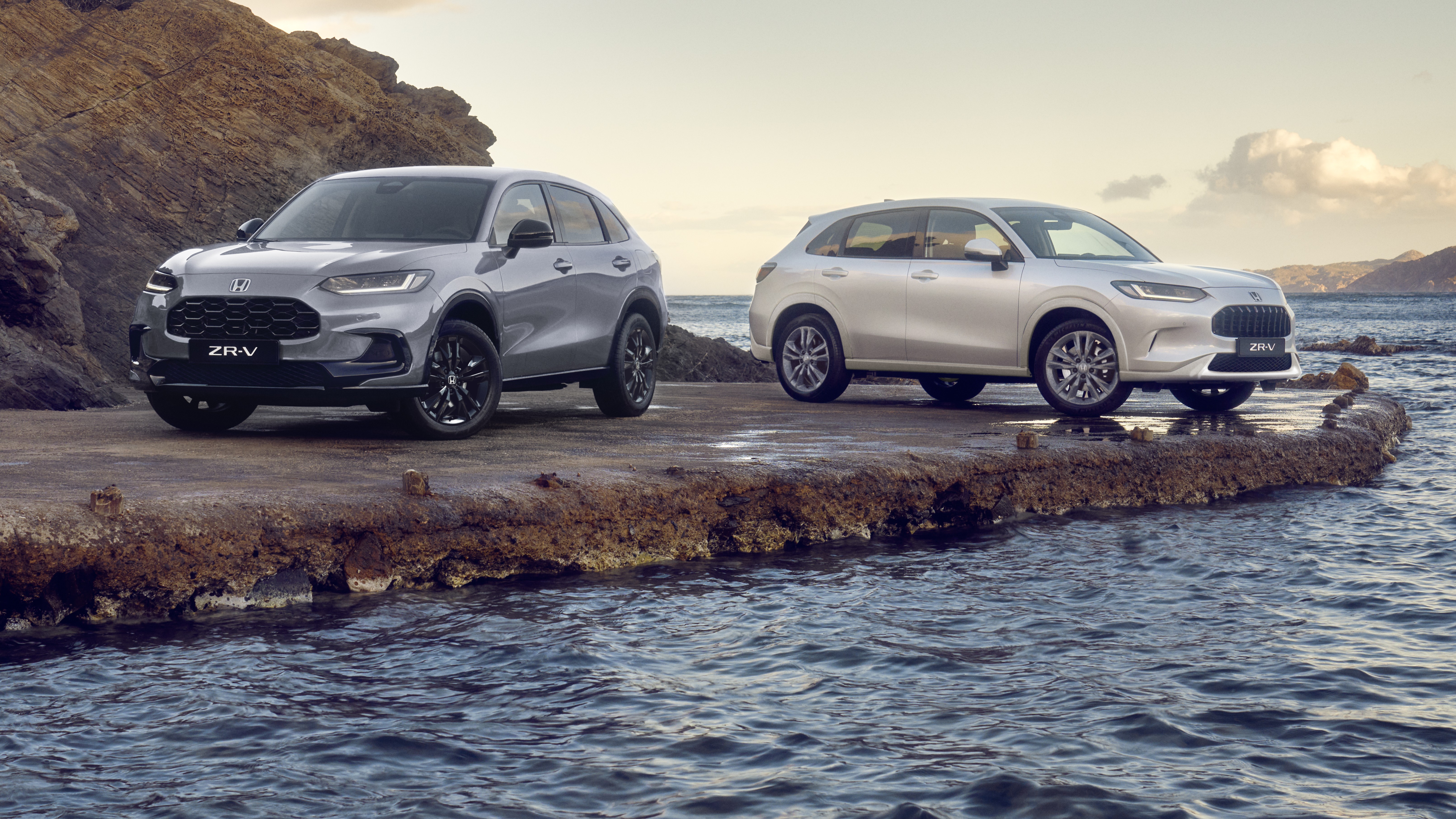 Two Honda ZR-V SUVs, one blue and one white, parked on a rocky coastal outcrop with water and hills in the background under a partly cloudy sky.