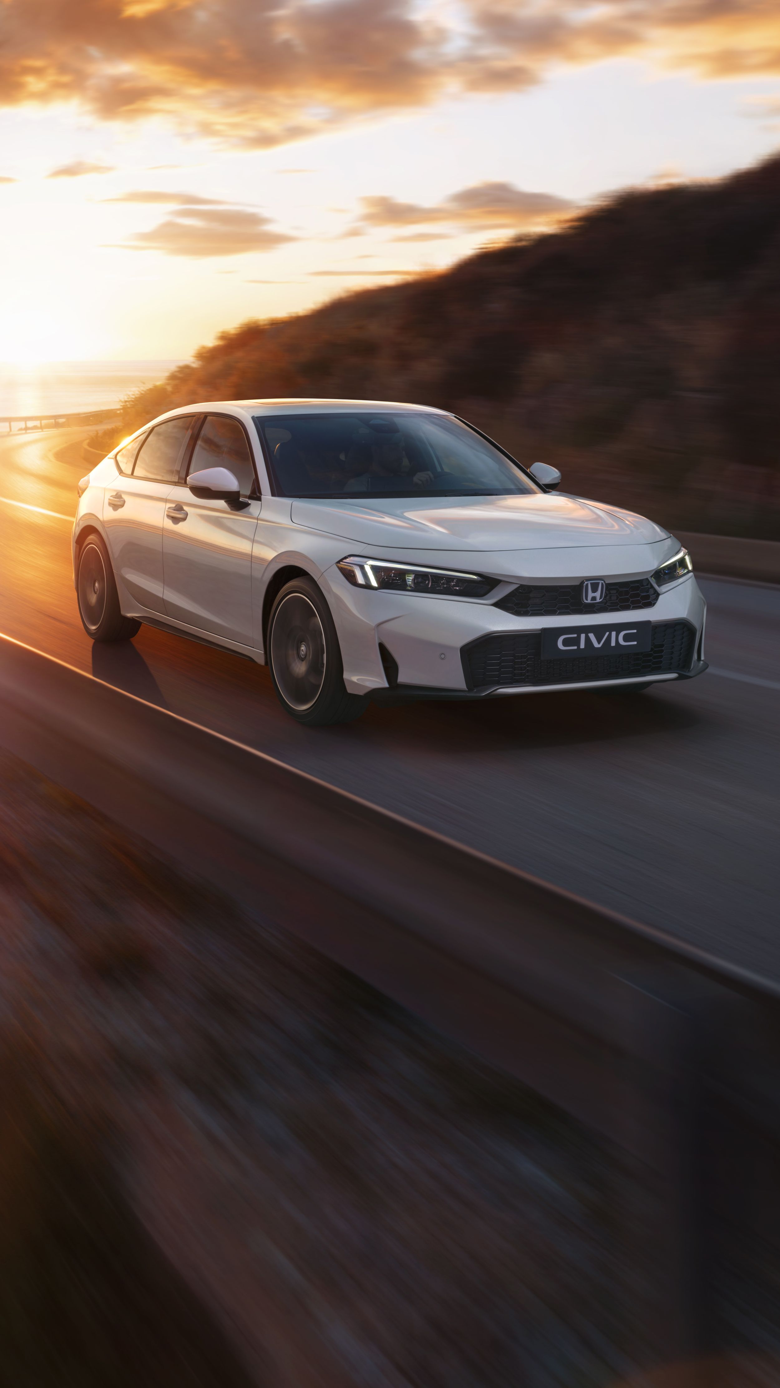 Egy fehér Civic Hybrid halad egy tengerparti úton naplementekor.