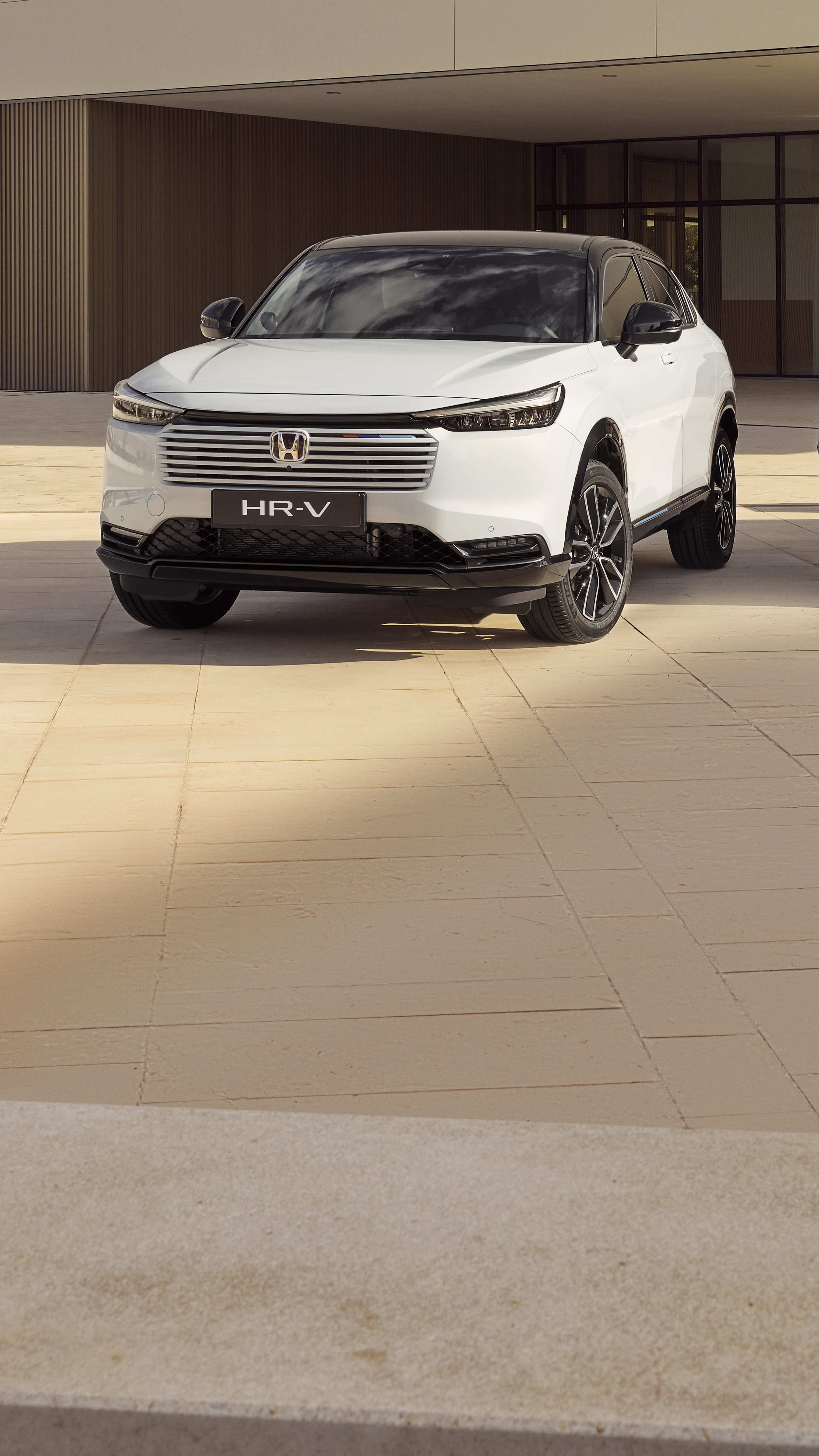 Egy fehér Honda HR-V SUV parkol egy modern, bézs csempézett falú épület előtt. A jármű kissé jobbra fordulva áll, bemutatva áramvonalas kialakítását és jellegzetes hűtőrácsát.