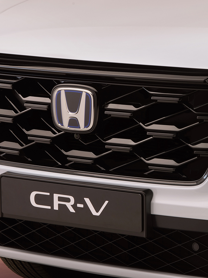Közelkép egy Honda CR-V első hűtőrácsáról, amelyen fekete, méhsejtmintás rács, középen a Honda embléma, alatta pedig a CR-V modellnév látható.