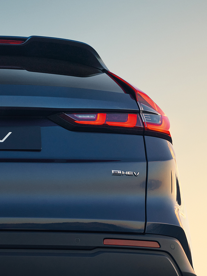 Egy sötétkék Honda CR-V e:HEV hátsó közelképe, amelyen a Honda jelvény, a CR-V felirat és a megvilágított, karakteres LED-es hátsó lámpák láthatók a lágy naplementés égbolt előtt.