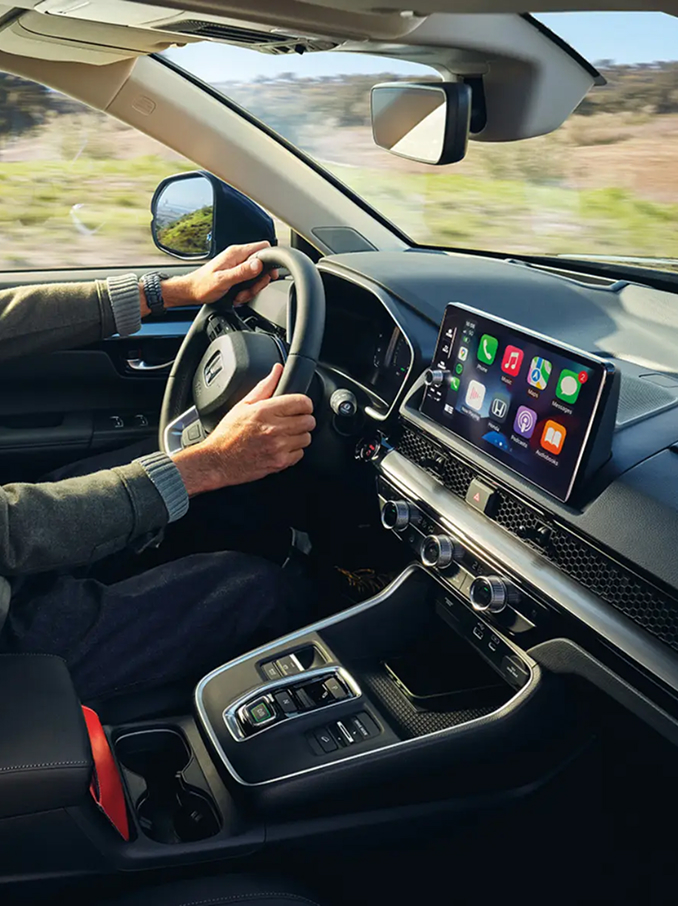Egy modern autó belső nézete, ahol a vezető a kormány mögött ül; látható a műszerfal, a kormánykerék, a középkonzol és egy széles érintőképernyős kijelző színes alkalmazásikonokkal, miközben az ablakokon kívül vidéki táj vonul el.