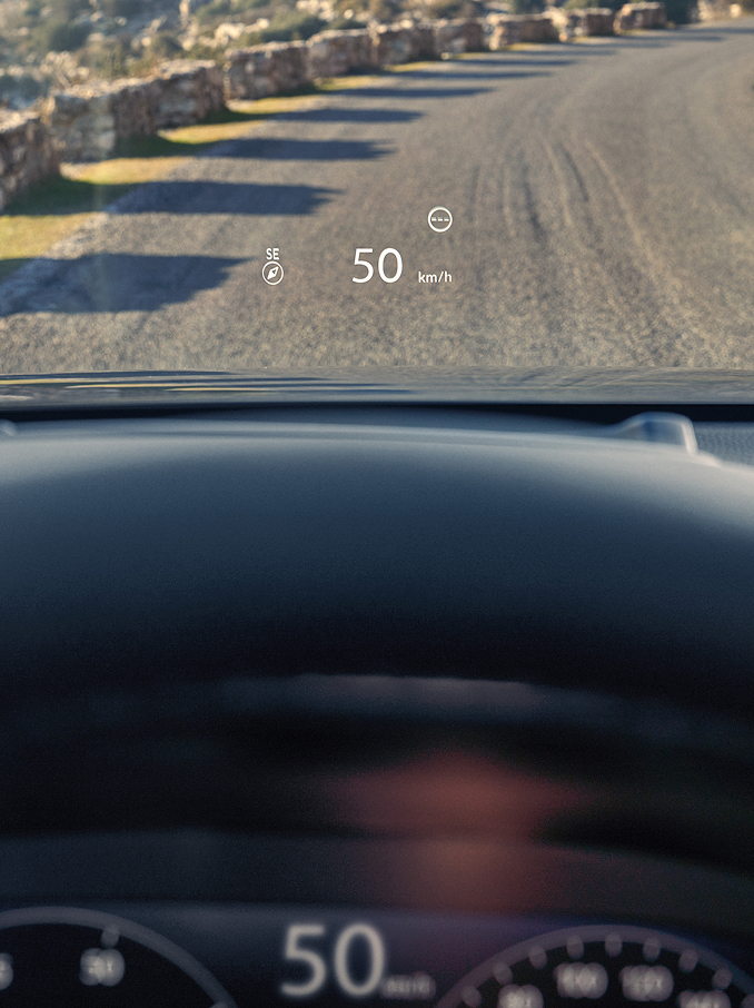 Első személyű nézet a vezetőülésből, ahol a Head-Up Display (HUD) a szélvédőre vetíti a „50 km/h” sebességet és egy iránytűt egy kavicsos út előtt.