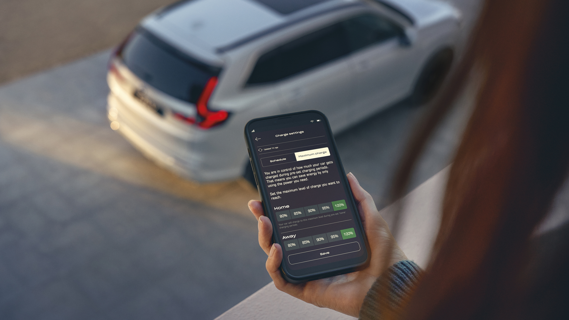 Egy személy egy okostelefont tart a kezében, amelyen egy járműtöltési beállításokat megjelenítő alkalmazás látható, miközben a háttérben egy fehér SUV parkol.