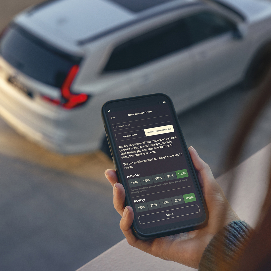 Egy személy egy okostelefont tart a kezében, amelyen egy járműtöltési beállításokat megjelenítő alkalmazás látható, miközben a háttérben egy fehér SUV parkol.