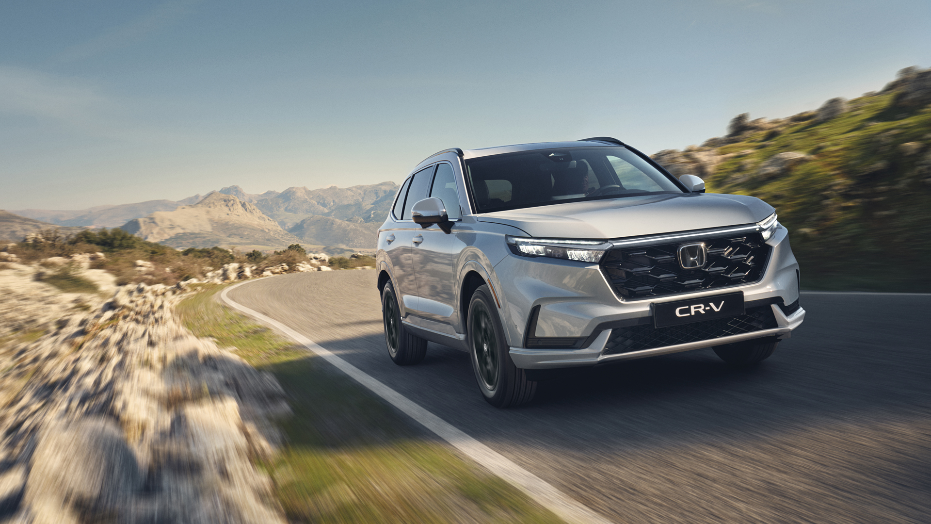 A silver Honda CR-V SUV driving on a winding mountain road with rocky terrain and mountains in the background. The vehicle is in motion, creating a sense of speed and adventure in the rugged landscape.