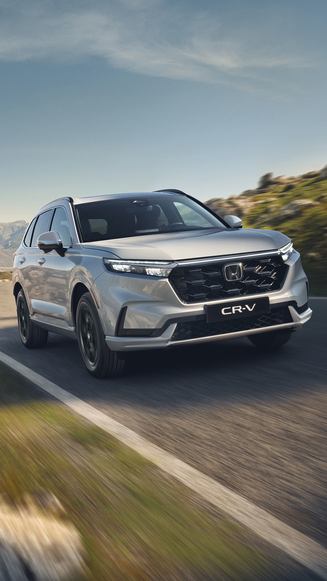 Egy ezüstszínű Honda CR-V SUV kanyargós hegyi úton halad, a háttérben sziklás terep és hegyek, tiszta kék ég alatt.