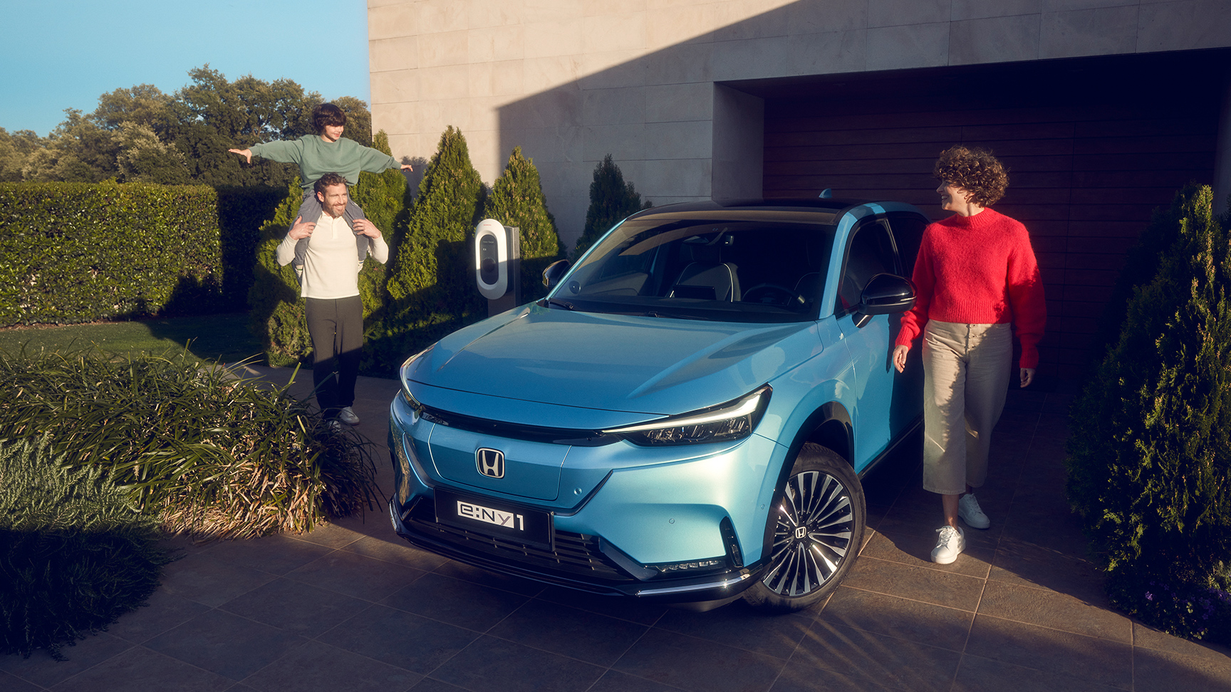 Egy ház előtt parkoló Honda e:Ny1 elölről, mellette egy család áll.