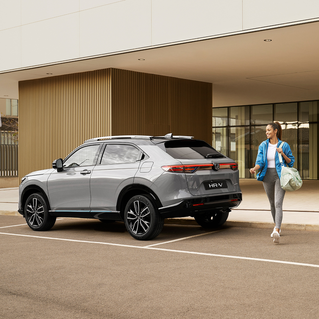 Egy ezüst Honda HR-V egy modern épület előtt parkol, közben egy nő sétál el mellette egy táskával, ami a mindennapi városi használatra és kényelemre utal.