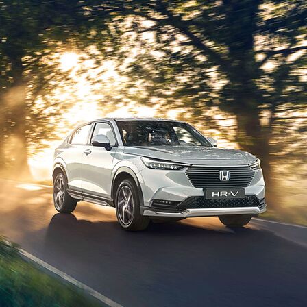 Háromnegyedes felvétel a Honda HR-V Hybrid elejéről, amely egy városon kívüli úton halad.