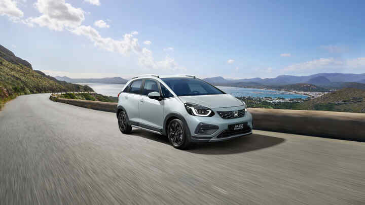 Elölről, háromnegyedes szögből készült felvétel a Honda Jazz Hybrid modellről, amelyet egy festői látkép előtt vezetnek.