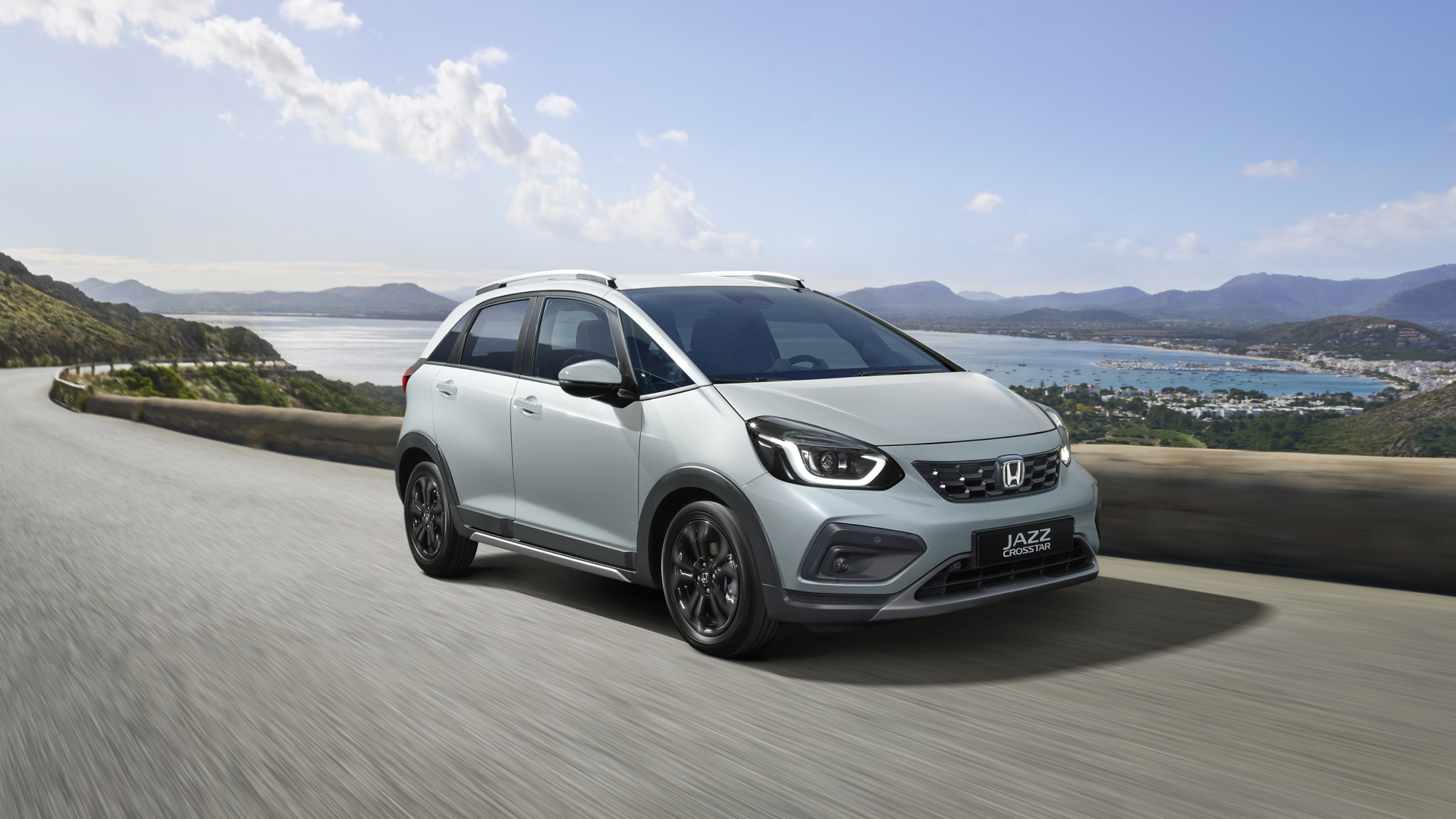 Elölről, háromnegyedes szögből készült felvétel a Honda Jazz Hybrid modellről, amelyet egy festői látkép előtt vezetnek.