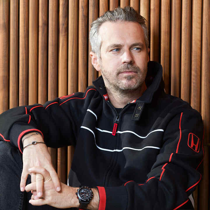 Egy Honda Racing sapkát és kabátot viselő férfi, aki a kalapja karimájához érinti az ujját.