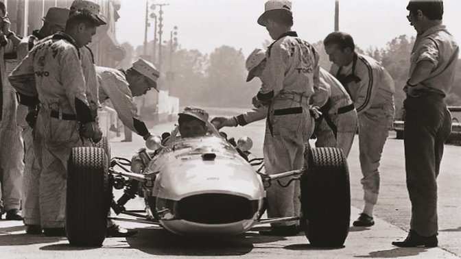 Egy hatvanas évek-beli, F1-es Honda versenyautó elölről, versenyzővel és mérnökökkel.