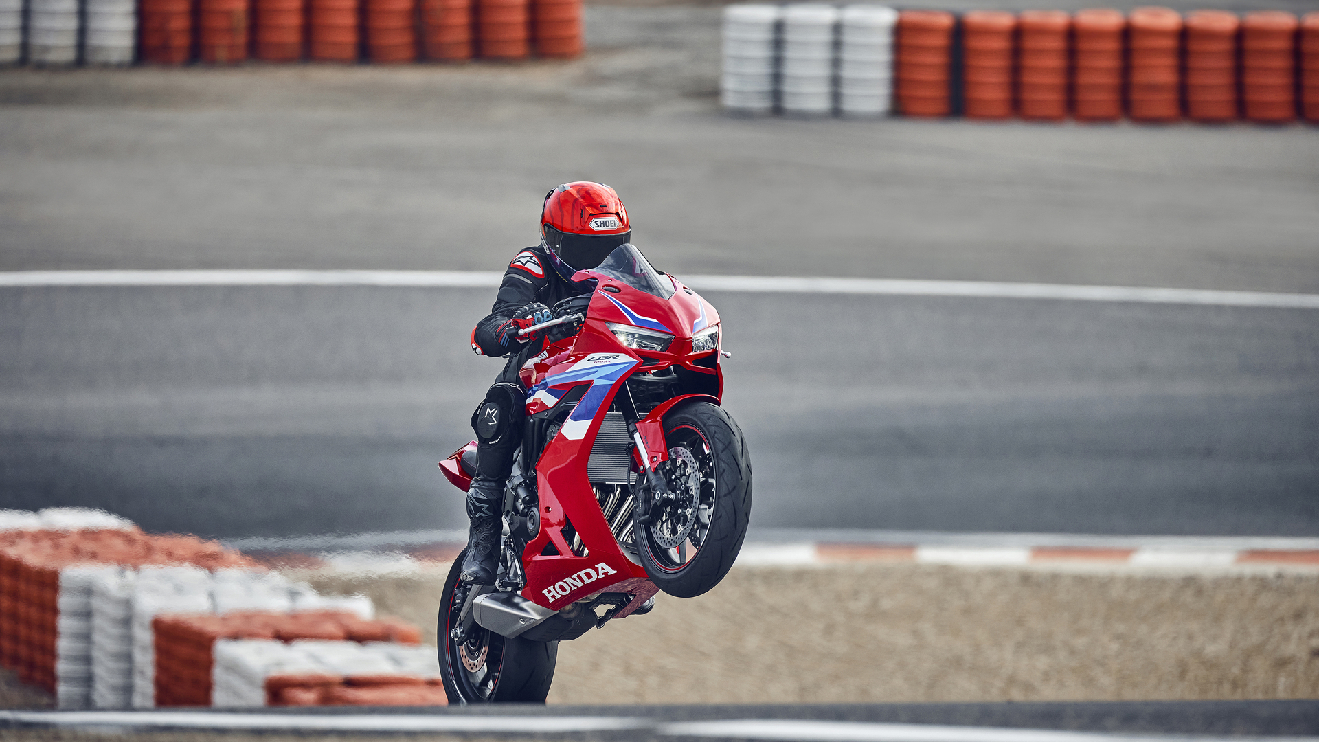 A red Honda CBR1000RR-R Fireblade motorcycle performing a wheelie on a race track, with orange and white safety barriers visible in the background.