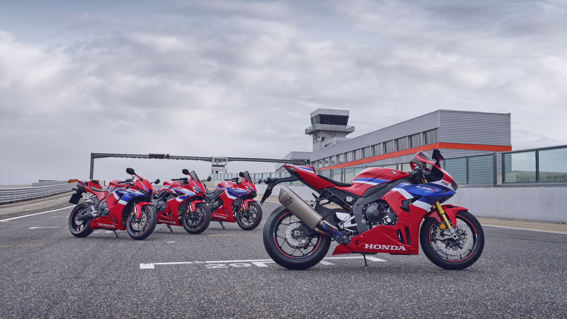 A lineup of five red Honda CBR sportbikes parked on a racetrack, showcasing different models from the series. The bikes are arranged in a row with an airport control tower visible in the background under an overcast sky.
