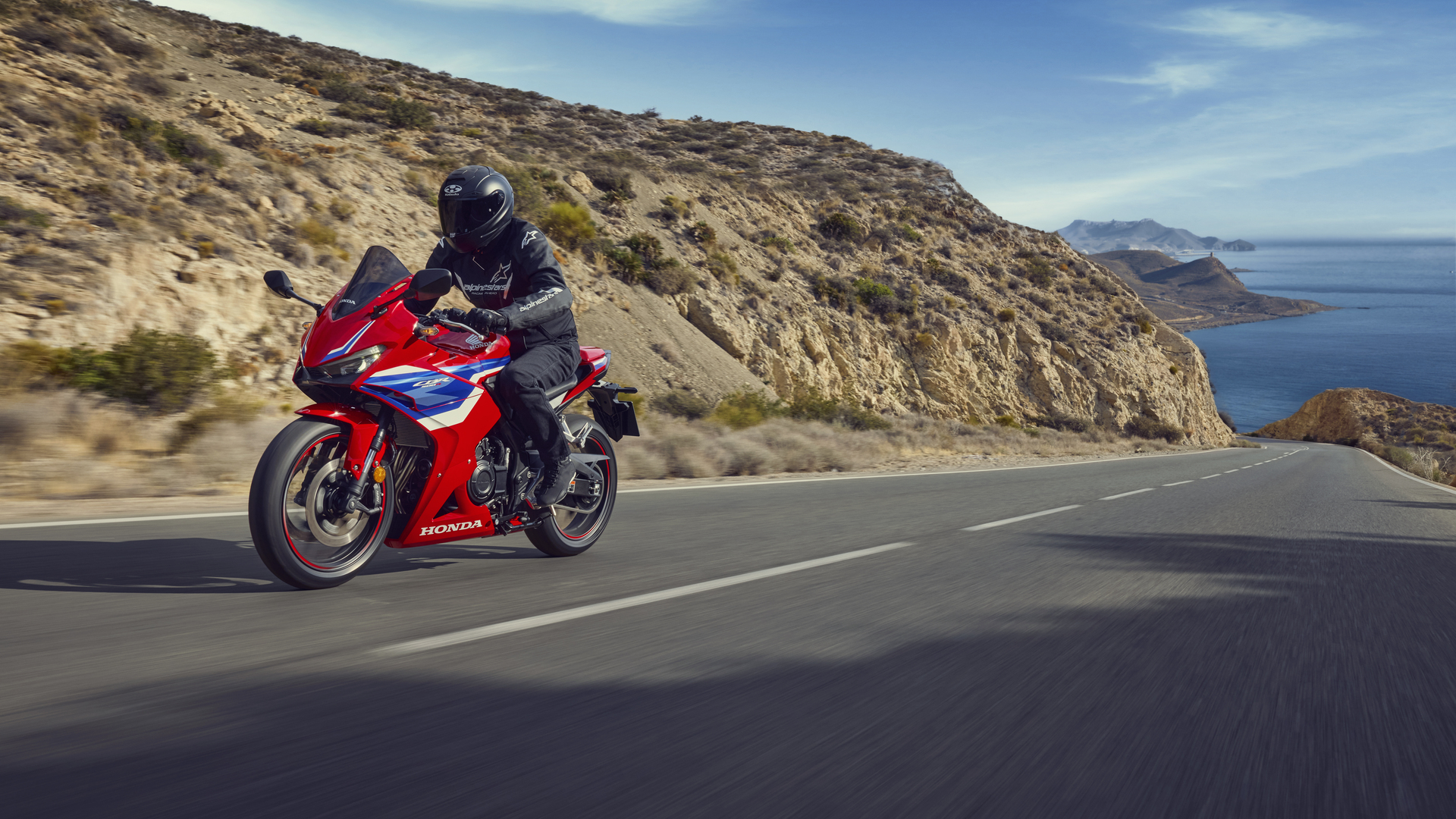 A red and blue Honda CBR650R motorcycle speeds along a coastal road, with a rider in black gear. The road curves around a rocky hillside, with the ocean visible in the background under a blue sky.