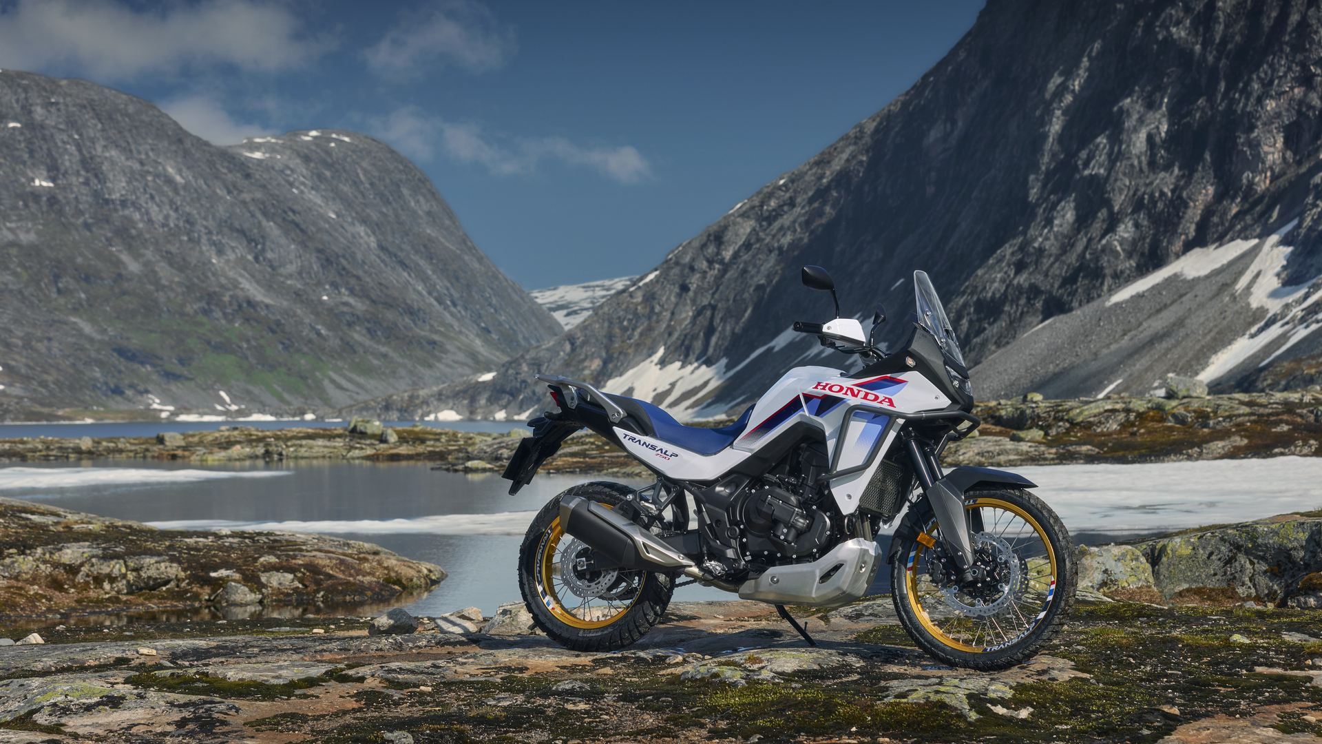 A Honda Africa Twin adventure motorcycle parked on rocky terrain beside a mountain lake, with snow-capped peaks in the background. The bike's white, blue, and red color scheme stands out against the rugged landscape.