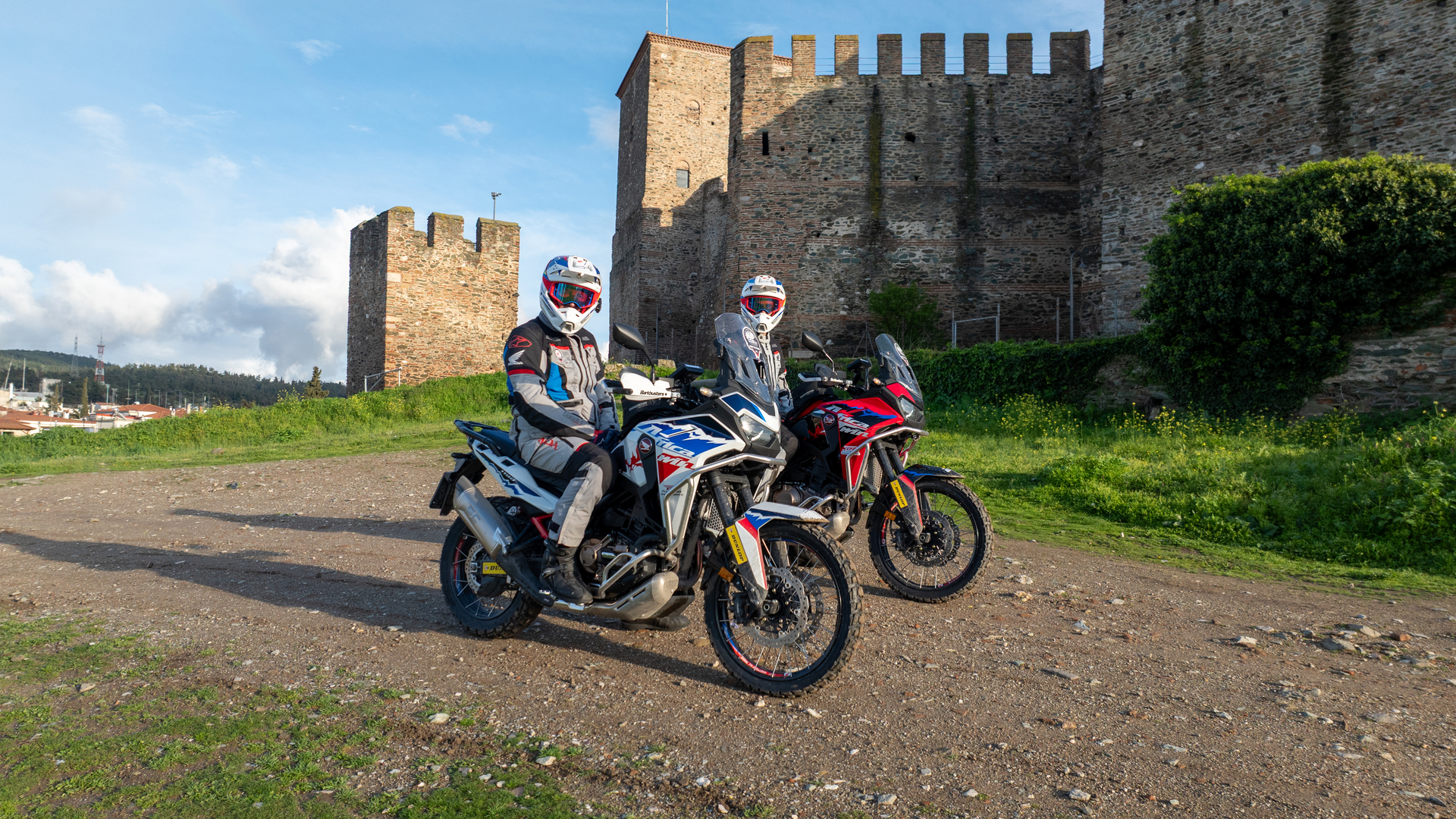 Two Honda Africa Twin adventure motorcycles parked in front of a medieval stone castle with crenellated walls. Riders in full gear are seated on the bikes, ready for an off-road journey.
