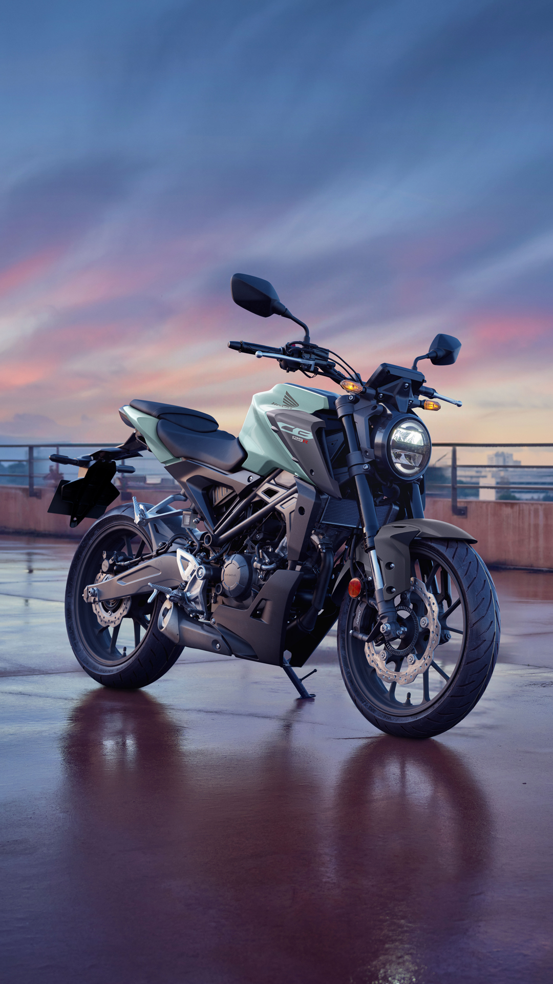 A Honda CB300R motorcycle parked on a rooftop at sunset. The bike is mint green and black, with a sleek, modern design. The sky above is filled with purple and pink hues, creating a dramatic backdrop for the motorcycle.