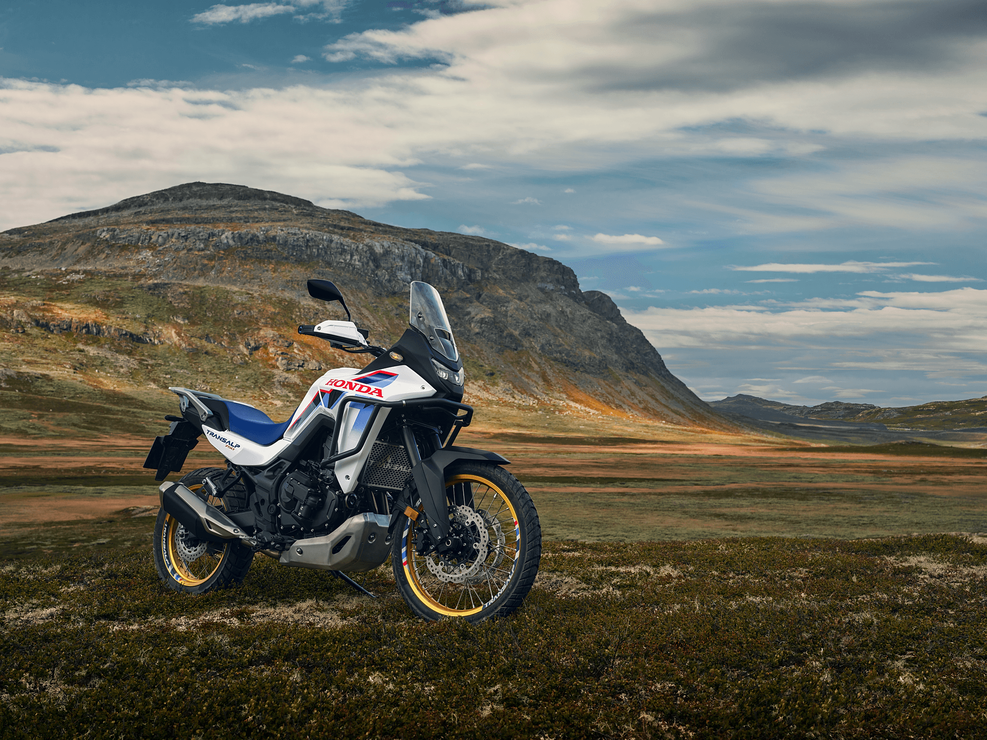 A Honda Africa Twin adventure motorcycle parked on rugged terrain with mountains in the background. The bike is white, blue, and red, featuring gold wheels and a tall windscreen. The landscape shows a grassy plain leading to rocky hills under a partly cloudy sky.