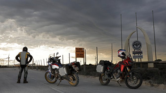 A horizontot kémlelő motoros, ahogy két, az aszfalton leállított Africa Twin mellett áll.