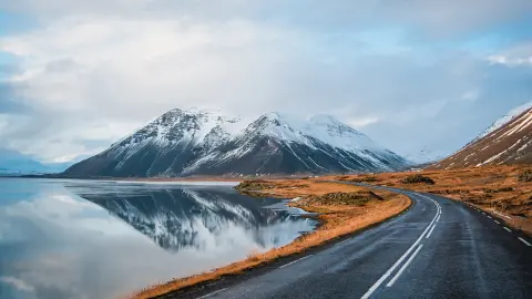 Téli panorámakép egy tó partja mentén vulkanikus hegyek felé vezető útról. Vízfelszínen tükröződő magas, sziklás, hófödte csúcsok. Az izlandi 1-es főútvonal a járművezető szempontjából.