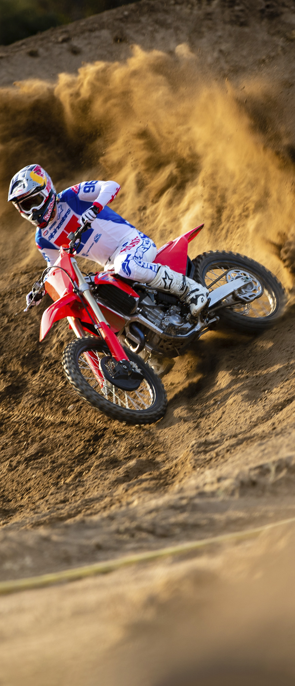 Egy fehér, kék és piros felszerelést viselő motocrossos egy piros terepmotoron bedől a kanyarban egy meredek, homokos pályán, közben nagy porfelhőt ver fel, ahogy nagy sebességgel áthajt a kanyaron.