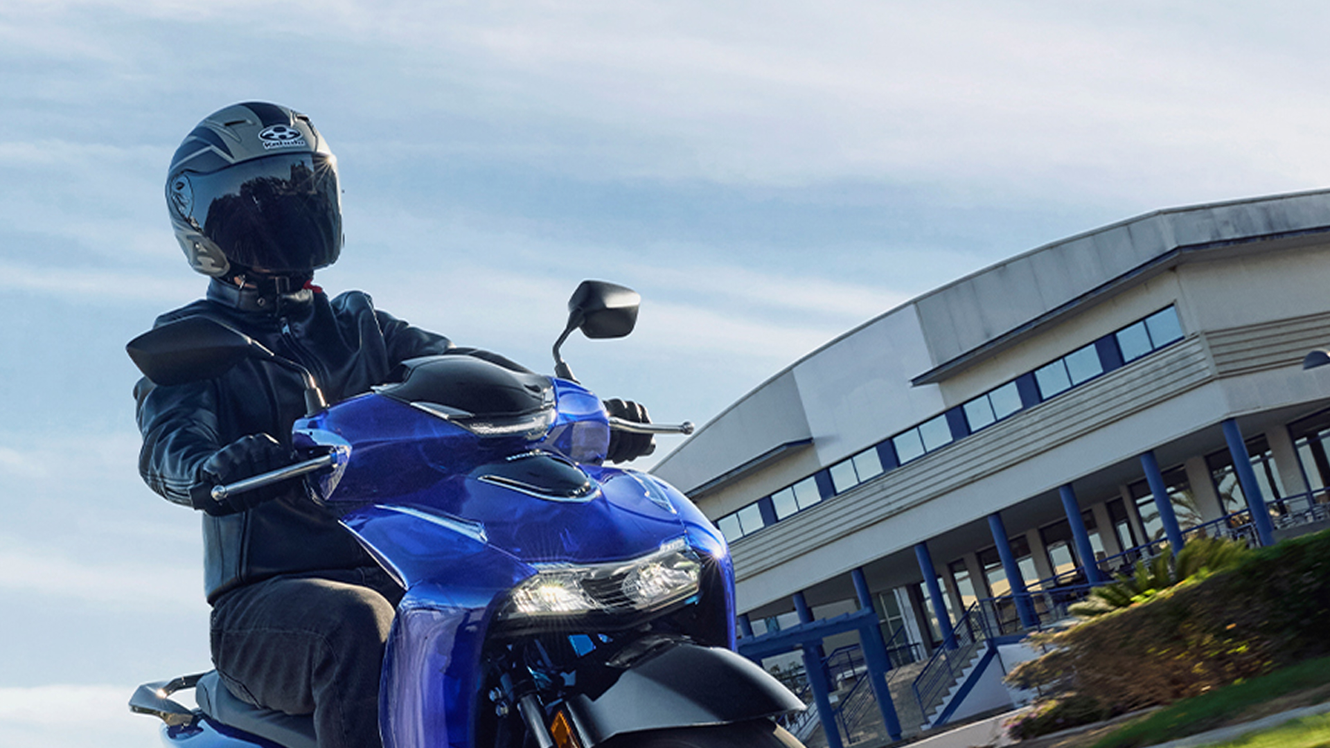 A person in black riding gear sits on a blue Honda sport motorcycle, likely a CBR model, in front of a modern building with curved architecture. The rider wears a black helmet and the motorcycle's front fairing and headlights are visible.