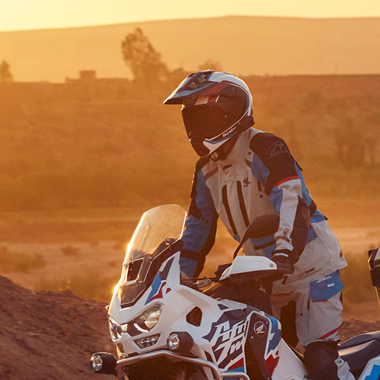 A motorcyclist riding a Honda Africa Twin adventure bike in a desert landscape at sunset. The rider is wearing protective gear and a helmet, silhouetted against the orange sky.