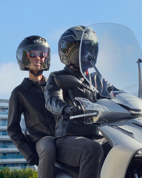 Two people sitting on a silver Honda scooter, likely a PCX model, wearing black helmets and clothing. The rider in front is gripping the handlebars, while the passenger sits behind. A clear windshield and blue sky are visible in the background.