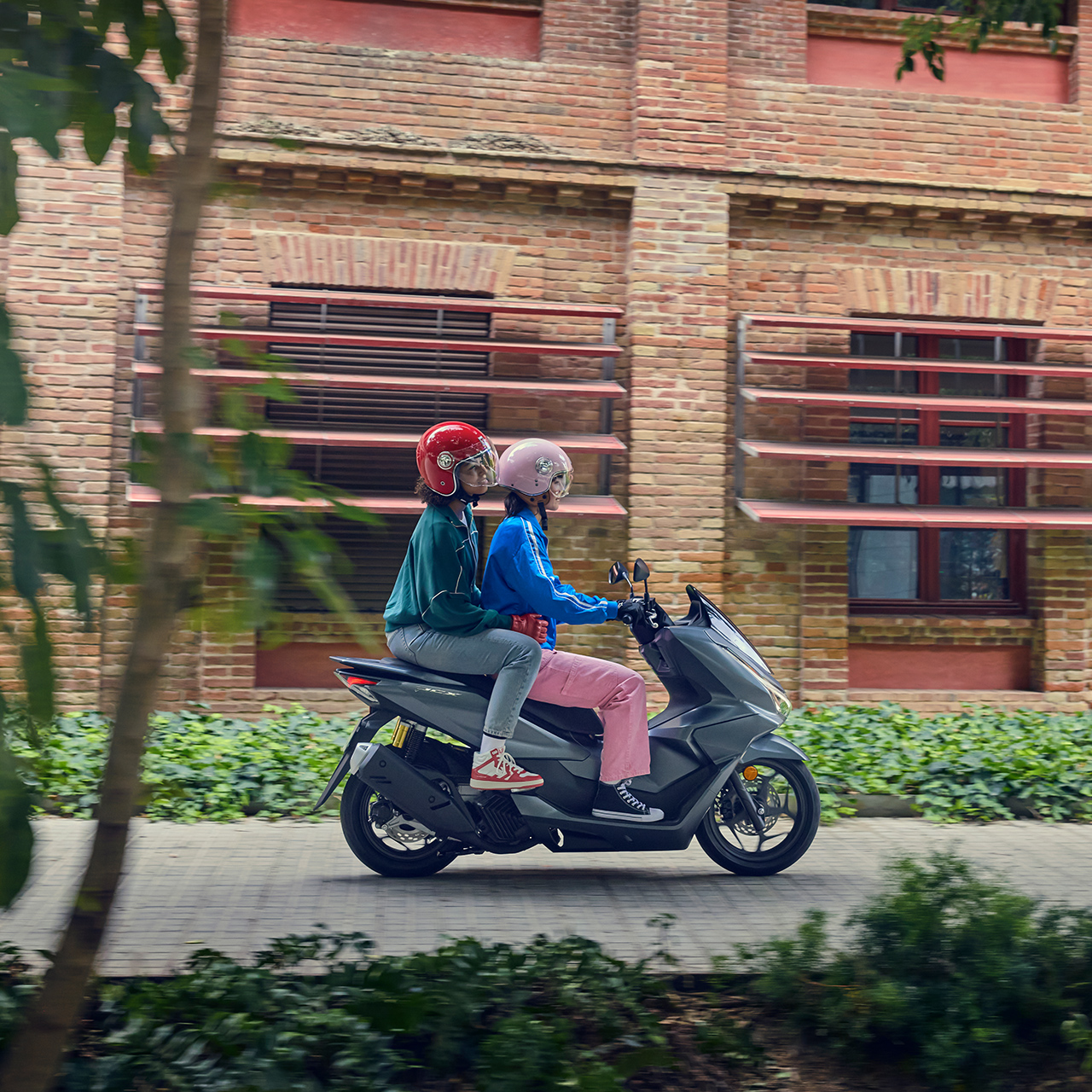 Oldalról készült felvétel a PCX125 motorkerékpáron utazó fotómodellekről egy városi helyszínen.