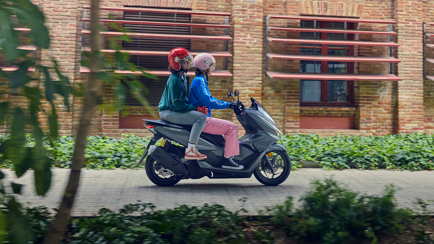 Oldalról készült felvétel a PCX125 motorkerékpáron utazó fotómodellekről egy városi helyszínen.