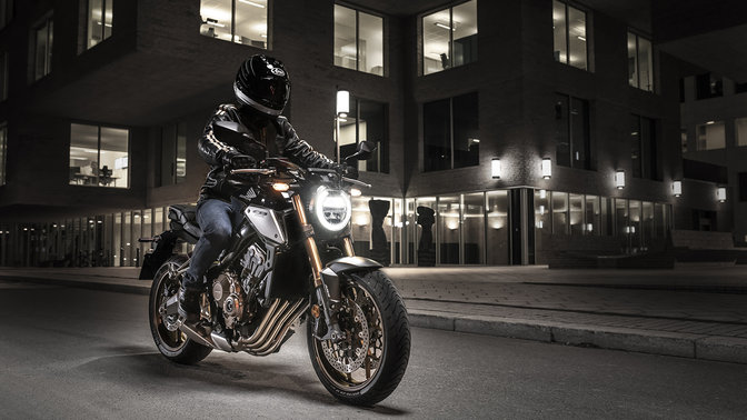 Háromnegyedes, elölről készült felvétel a CB650R Neo Sports Café jobb oldaláról pilótával városi környezetben éjszaka