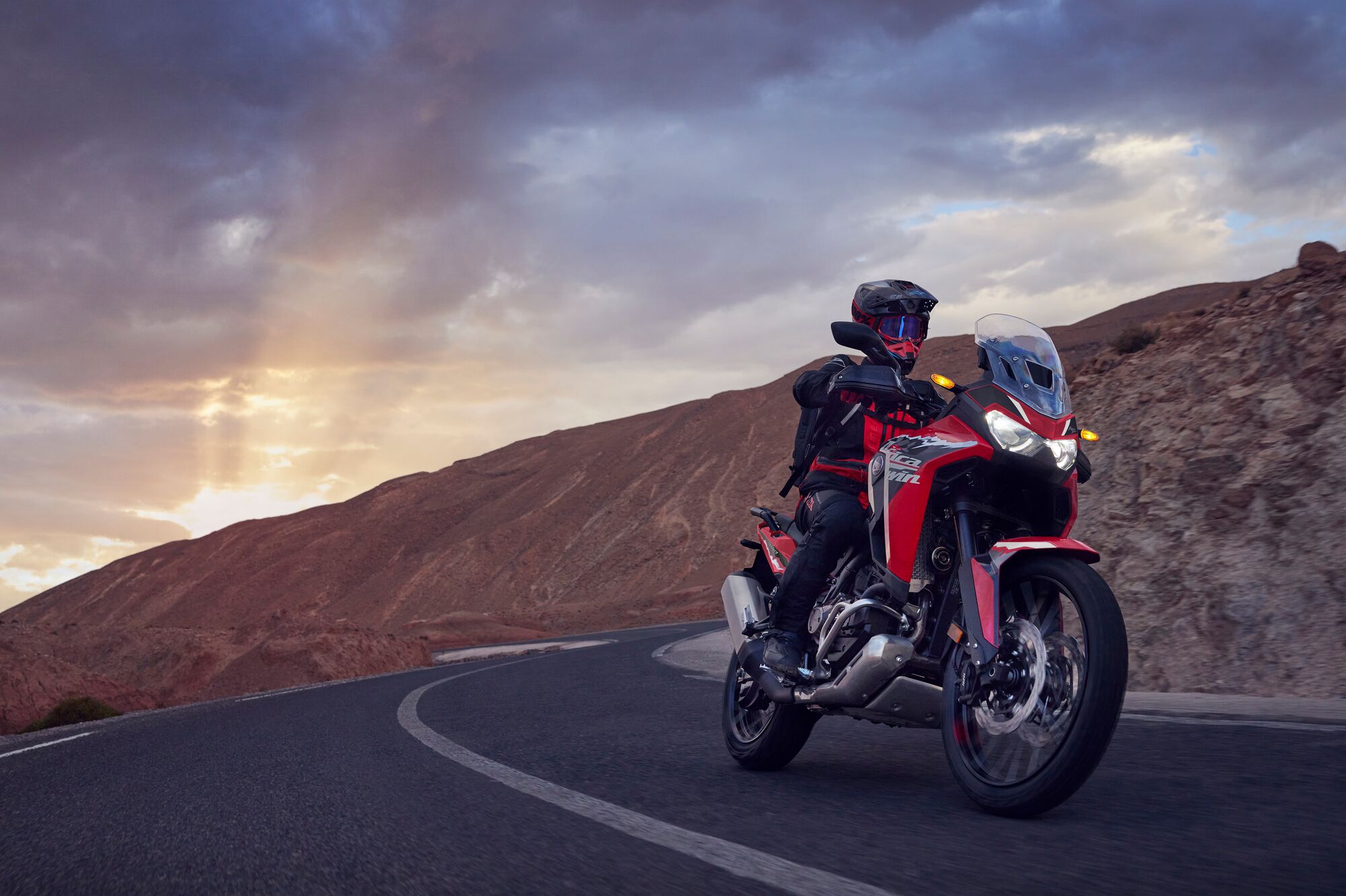A Honda Africa Twin motorcycle rides along a winding mountain road at sunset. The red and black adventure bike is navigating a curve with rocky terrain in the background under a dramatic, cloudy sky.
