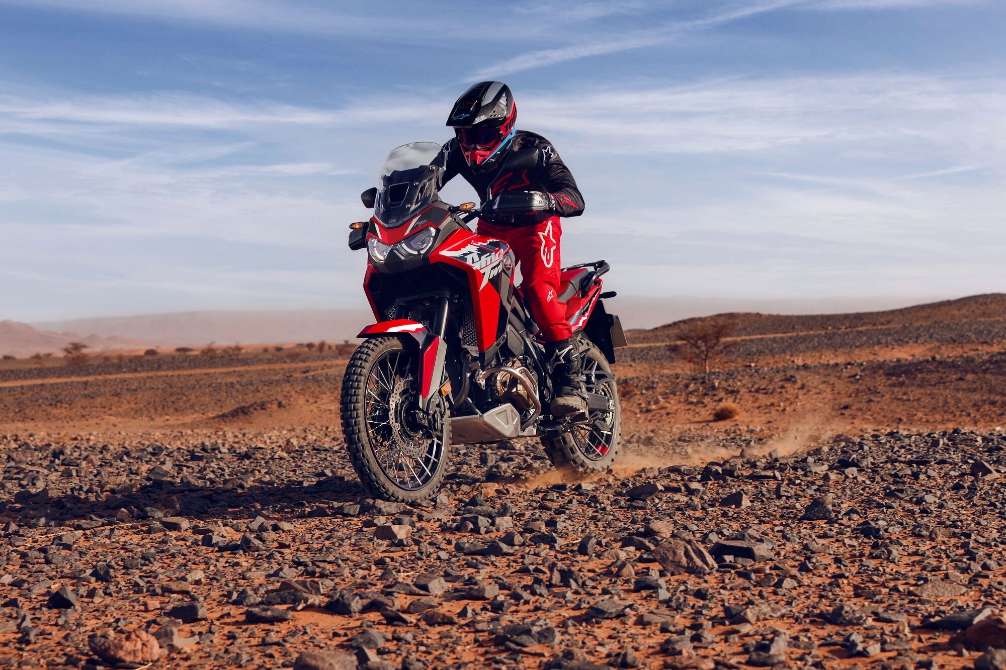 A red Honda Africa Twin motorcycle being ridden across a rocky desert landscape under a blue sky with wispy clouds.