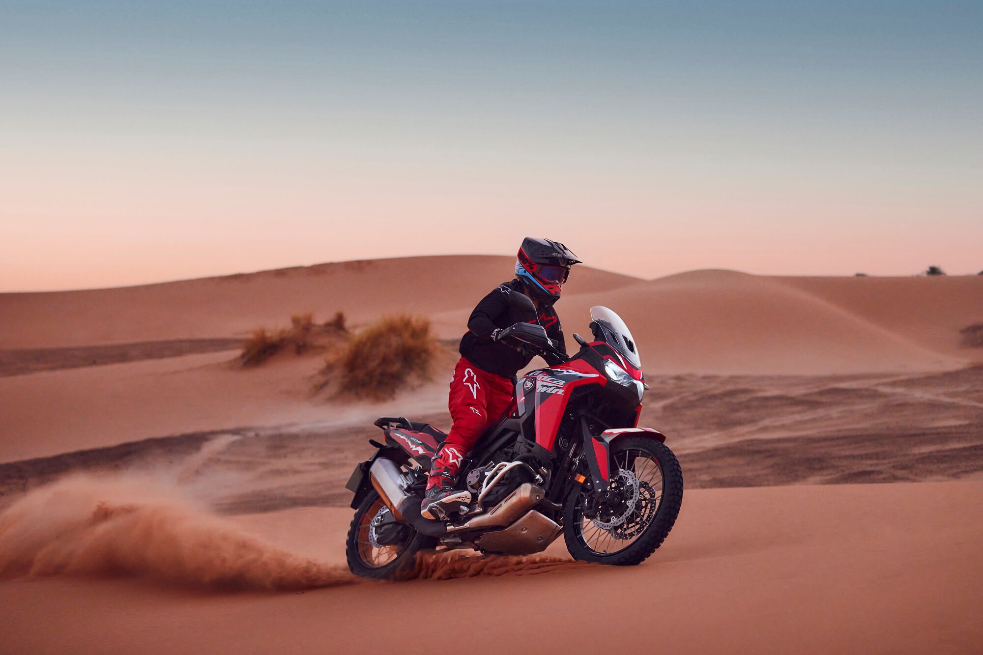 A rider on a red Honda Africa Twin motorcycle navigates through sandy desert dunes at sunset, kicking up a cloud of dust behind the bike.