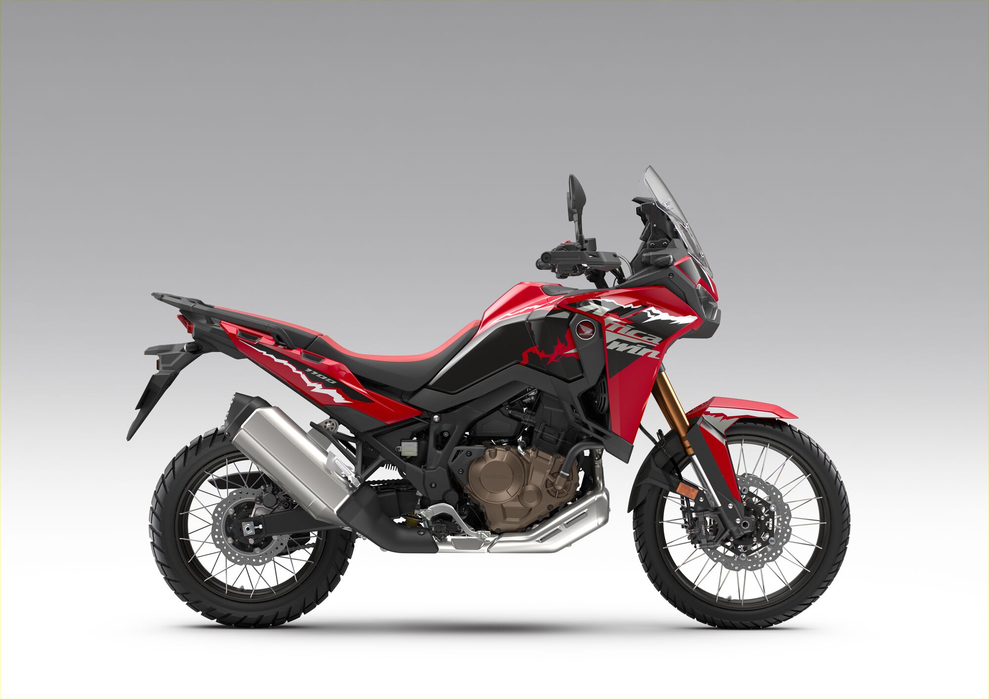 A side view of a Honda Africa Twin adventure motorcycle in red and black color scheme, featuring a tall windscreen, rugged tires, and an upright riding position against a neutral gray background.