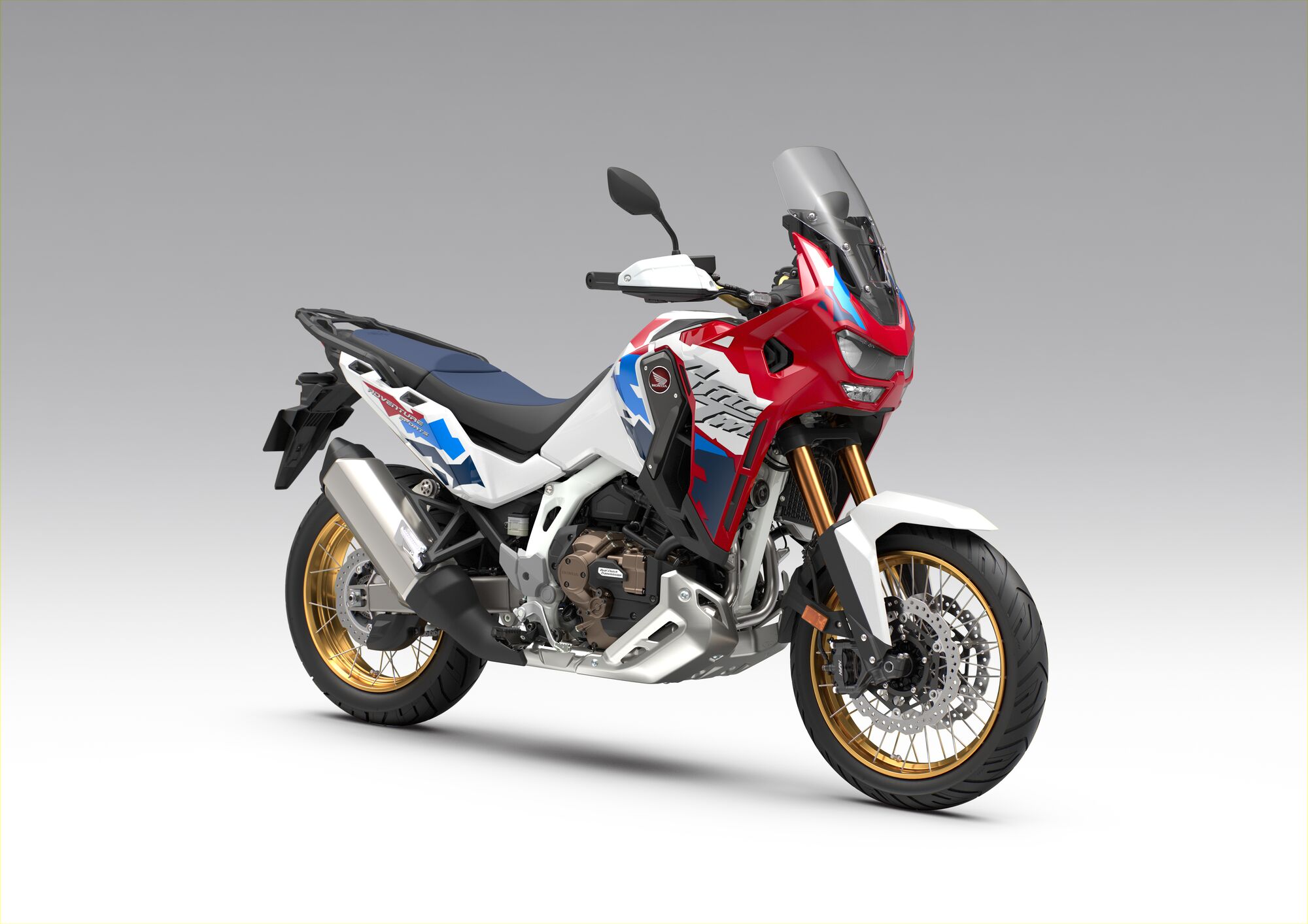 A Honda Africa Twin adventure motorcycle in red, white, and blue color scheme with gold wheels, featuring a tall windscreen and rugged styling, positioned against a plain gray background.