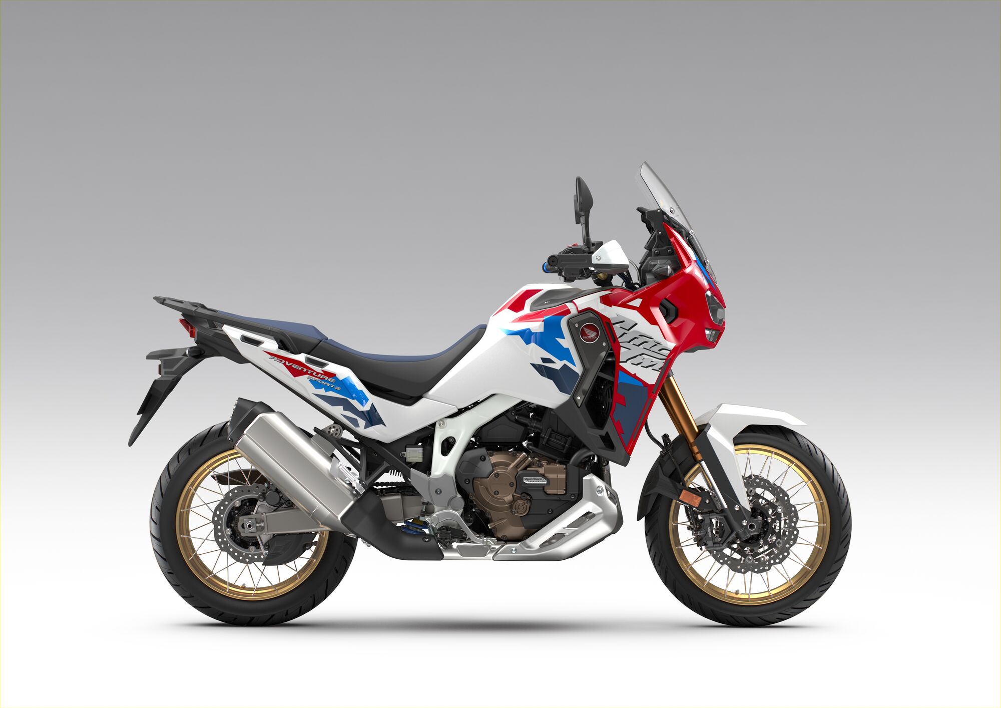A Honda Africa Twin adventure motorcycle in white, red, and blue color scheme with gold wheels, viewed from the side against a neutral background.