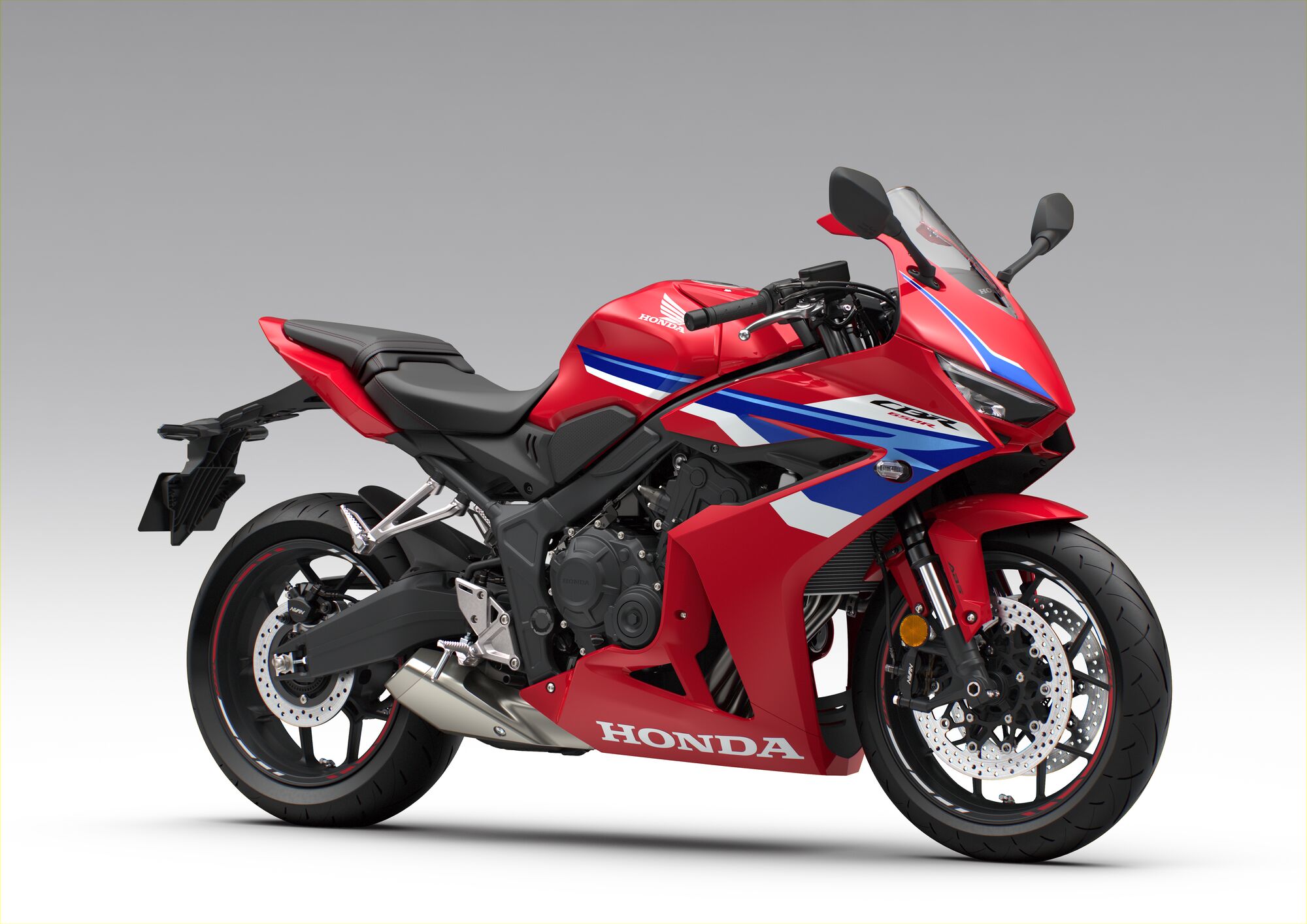 A red, blue, and white Honda CBR650R sport motorcycle viewed from the side against a neutral gray background. The sleek, aerodynamic design features sharp angles and a full fairing with the Honda logo prominently displayed.